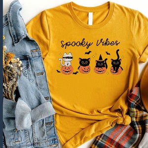 May include: A mustard yellow t-shirt with the text "Spooky Vibes" printed on the front. The shirt features a graphic of four black cats sitting in pumpkins.