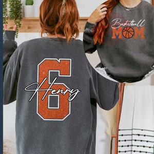 May include: A gray sweatshirt with an orange glitter number 6 and the name "Henry" in white script. The sweatshirt is shown on a model from the back. A second gray sweatshirt is shown on a model from the front with the text "Basketball Mom" and a basketball graphic in orange glitter.