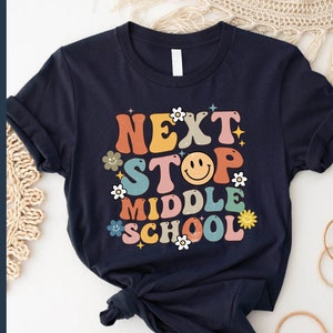 May include: A dark blue t-shirt with the text "Next Stop Middle School" in colourful retro lettering. The text is surrounded by flowers and a smiley face.