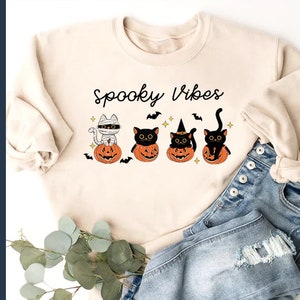 May include: A beige sweatshirt with the text "Spooky Vibes" and a graphic of four black cats sitting in pumpkins. The cats are dressed in Halloween costumes, including a mummy, a witch, and a cat.