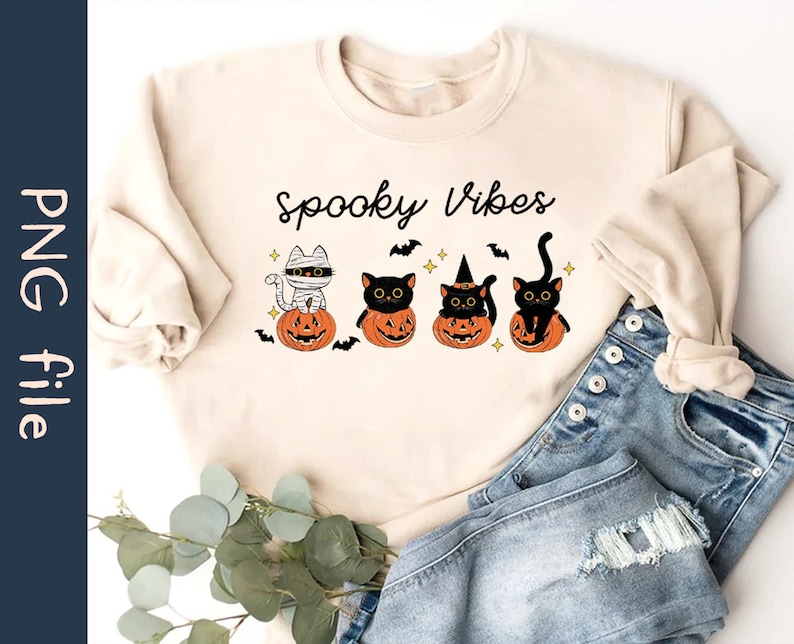 May include: A beige sweatshirt with the text "Spooky Vibes" and a graphic of four black cats sitting in pumpkins. The cats are dressed in Halloween costumes, including a mummy, a witch, and a cat.
