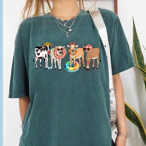 May include: A teal blue t-shirt with a graphic of five cows wearing sunglasses and floral headbands. The cows are holding onto a colorful inflatable ring.