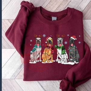 May include: A maroon sweatshirt with a Christmas-themed design featuring four Great Danes wearing festive attire. The dogs are wearing Santa hats, reindeer antlers, and Christmas lights. The design is centered on the sweatshirt.