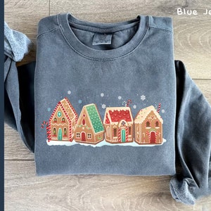 May include: A blue sweatshirt with a graphic of five gingerbread houses with red and white candy canes, red and white candy hearts, and white snowflakes. The houses are decorated with red and white candy.