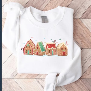 May include: White sweatshirt with a graphic of gingerbread houses with red and green accents. The houses are decorated with candy canes and snowflakes.