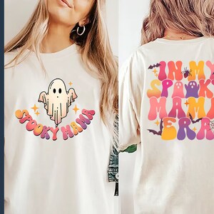 Op de afbeelding: Twee witte T-shirts met een Halloween-thema. E&eacute;n shirt heeft een cartoon-geest met de tekst "Spooky Mama" in regenboogkleuren. Het andere shirt heeft de tekst "In My Spooky Mama Era" in regenboogkleuren met een spin, vleermuizen en een spinnenweb.