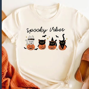 May include: A white t-shirt with the text "Spooky Vibes" printed on it. There are four black cats in the image, each sitting in a jack-o'-lantern. One cat is wearing a mummy costume, another is wearing a witch's hat, and the other two are wearing nothing.