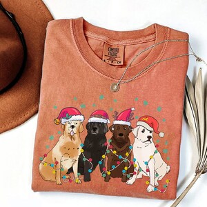 Puede incluir: Una camiseta roja, naranja y blanca con un dise&ntilde;o navide&ntilde;o que presenta cuatro perros con gorros de Pap&aacute; Noel y rodeados de luces navide&ntilde;as de colores.