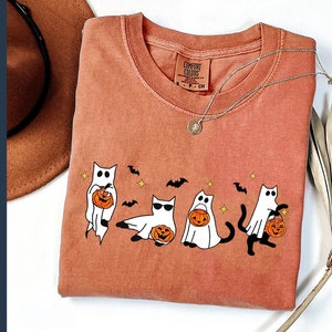 May include: Rust-colored Comfort Colors t-shirt featuring a Halloween-themed graphic. The design includes cartoon cats dressed as ghosts, holding jack-o'-lanterns. The shirt also has bats and stars. A silver necklace with a pendant is on the shirt.