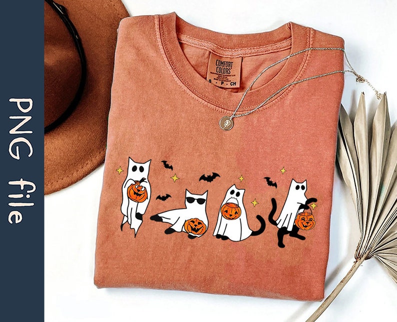 May include: Rust-colored Comfort Colors t-shirt featuring a Halloween-themed graphic. The design includes cartoon cats dressed as ghosts, holding jack-o'-lanterns. The shirt also has bats and stars. A silver necklace with a pendant is on the shirt.
