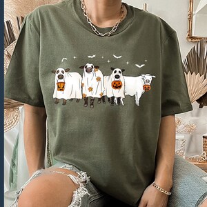 May include: A green t-shirt with a graphic of four cows dressed as ghosts holding buckets of candy. The cows are wearing white sheets with black eyes and mouths. The text "Boo" is printed on the shirt.