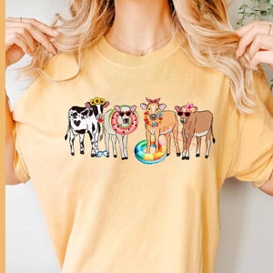 May include: A yellow t-shirt with a graphic of four cows wearing flower crowns and holding pool floats. The cows are black and white, brown and white, and brown.