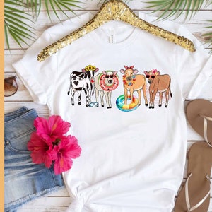 May include: White t-shirt with a graphic of four cows wearing sunglasses and summer accessories. The cows are wearing flower crowns, sunglasses, and inflatable pool rings.