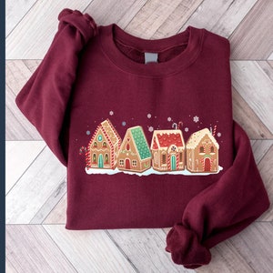 May include: A maroon sweatshirt with a graphic of four gingerbread houses with white frosting and candy decorations. The houses are arranged in a row and have green, red, and white accents.