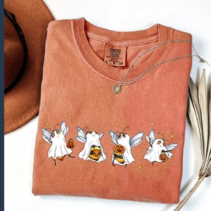 May include: A burnt orange t-shirt with a graphic of five cartoon ghosts holding jack-o-lanterns. The ghosts are wearing bee wings and have a cute, whimsical look. The text "COMFORT COLORS" is visible on the shirt.