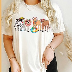 May include: A white t-shirt with a graphic of five cows wearing sunglasses and floral headbands. The cows are standing in a line and one is holding a rainbow float.
