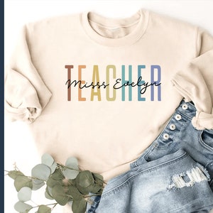 Puede incluir: Una sudadera beige con el texto "TEACHER" en colores arco&iacute;ris y el texto "Miss Evelyn" en letra cursiva negra.