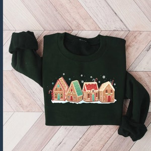 May include: A dark green sweatshirt with a festive design of gingerbread houses with white frosting and candy canes. The houses are arranged in a row and have red, green, and white accents.