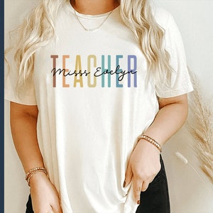 Puede incluir: Una camiseta blanca con el texto "TEACHER" en colores arco&iacute;ris y el texto "Miss Evelyn" en letra cursiva negra.