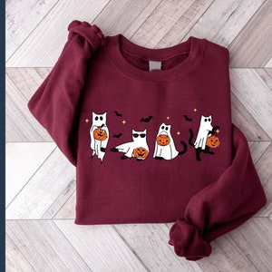 May include: A maroon sweatshirt featuring a Halloween-themed design. The graphic depicts five cartoon cats dressed as ghosts, each holding a jack-o'-lantern. Bats and stars are also included in the design. The words "PNG file" are on the left side of the image.