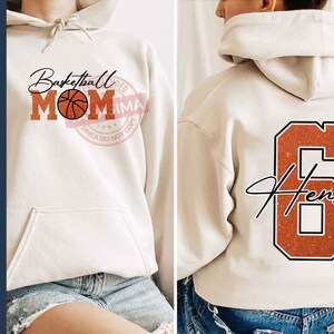 May include: A white hoodie with the text "Basketball Mom" and a basketball graphic on the front. The back of the hoodie has a large orange number 6 with the name "Henry" written in black.