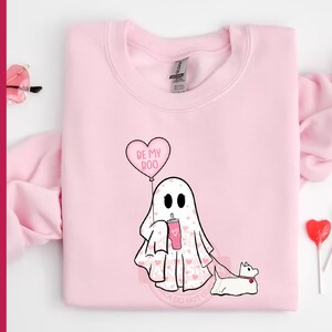 Puede incluir: Sudadera rosa con un fantasma que sostiene una bebida rosa y un globo en forma de coraz&oacute;n que dice "Be My Boo". El fantasma est&aacute; paseando a un peque&ntilde;o perro blanco.