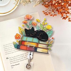May include: A badge reel featuring a black cat sleeping on a stack of books, surrounded by colorful flowers and leaves. The name "Tiffany" is written in cursive. The reel has a clear plastic retractable cord and a metal clip.