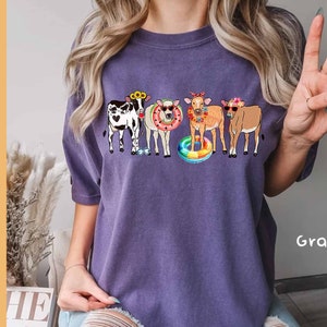 May include: Purple t-shirt with a graphic of four cows wearing sunglasses and floaties. The cows are wearing flowers in their hair. The text "Grape" is visible in the bottom right corner of the image.