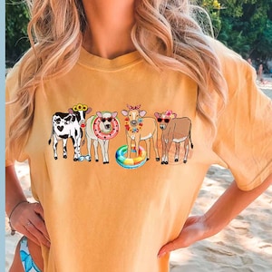May include: A yellow t-shirt with a graphic of four cows wearing sunglasses and flower crowns. The cows are standing in a line and one is holding a rainbow float.