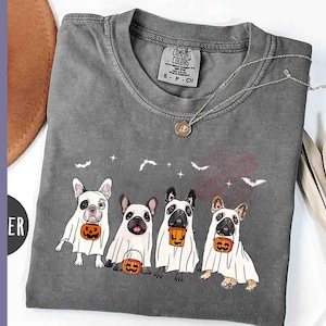 Pu&ograve; includere: Una maglietta grigia con una grafica di quattro bulldog francesi vestiti da fantasmi che tengono zucche. Il testo "Comfort Colors" &egrave; stampato sulla maglietta.