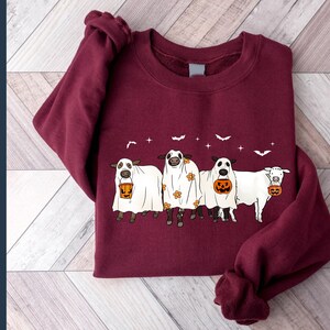 May include: A maroon sweatshirt with a graphic of four cows dressed as ghosts holding buckets of candy. The cows are wearing white sheets with floral patterns and have orange pumpkins in their buckets.