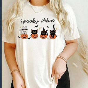 May include: White t-shirt with the text "Spooky Vibes" and a graphic of four black cats in pumpkins. The cats are wearing witch hats, a mummy costume, and one is just a regular black cat.