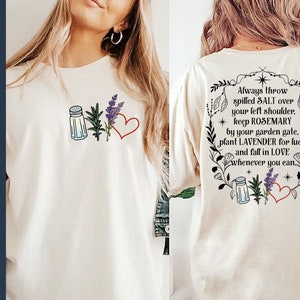 May include: A white t-shirt with a graphic design featuring a salt shaker, rosemary, lavender, and a heart. The text on the back of the shirt reads "Always throw spilled SALT over your left shoulder, keep ROSEMARY by your garden gate, plant LAVENDER for luck and fall in LOVE whenever you can."