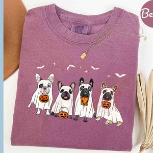 Pu&ograve; includere: Una maglietta viola con una grafica di quattro bulldog francesi vestiti da fantasmi per Halloween. Ogni cane tiene una piccola zucca. Il testo "Comfort Colors" &egrave; visibile sulla maglietta.