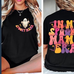 Op de afbeelding: Zwart T-shirt met een kleurrijke "Spooky Mama" afbeelding op de voorkant en "In My Spooky Mama Era" op de achterkant. De afbeelding bevat een spook en vleermuizen.