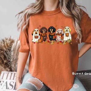 May include: A burnt orange t-shirt with a graphic of five dachshund dogs dressed in Halloween costumes. The dogs are wearing ghost, pumpkin, witch, and vampire costumes. The text "Burnt Orange" is visible in the bottom right corner of the image.