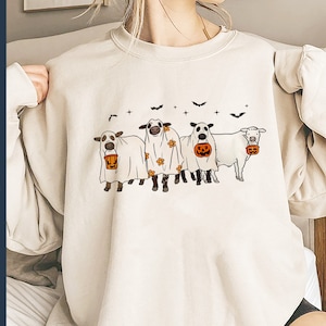 May include: A white sweatshirt with a graphic of four cows dressed as ghosts holding Halloween buckets. The cows are wearing white sheets with black eyes and mouths. The buckets are orange with a jack-o-lantern face.
