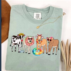 May include: A light green t-shirt with a graphic of four cows wearing sunglasses, flower crowns, and holding pool floats. The text "Comfort Colors" is visible on the shirt.