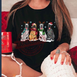 May include: A black t-shirt with a Christmas-themed design featuring four Great Danes wearing Santa hats and reindeer antlers. The dogs are wearing festive lights and are sitting in a row.