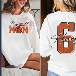 May include: White sweatshirt with the text "Basketball Mom" and a basketball graphic on the front. The back of the sweatshirt has a large orange number 6 with the name "Henry" written in cursive.