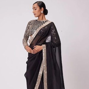 May include: A black sheer saree with a silver and gold mirror work border. The saree is draped on a person and has a sheer, flowing fabric.