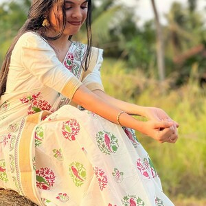 May include: A woman wearing a white saree with a floral pattern in red, green, and pink. The saree has a gold border and is draped elegantly. The background is a blurred outdoor scene with palm trees and greenery.