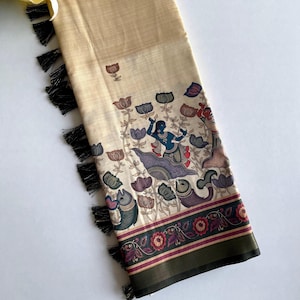 May include: A cream-colored silk saree with a black tassel border. The saree features a colorful floral and fish design with a blue figure in the center. The saree has a green and red border with a floral pattern.