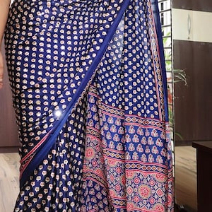 May include: A navy blue saree with a repeating white and gold pattern. The border features a red and gold design. The saree is draped, showcasing the fabric's texture and design. The saree has the text "DE" and "MRR-GJI-020225".