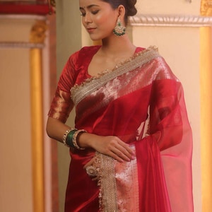 Pode incluir: Uma mulher usando um sari vermelho vibrante com detalhes dourados e uma blusa combinando. O sari apresenta uma borda prateada brilhante e é combinado com pulseiras e brincos verdes. A aparência geral é elegante e tradicional.