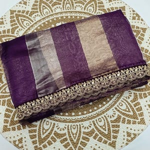 May include: A folded purple and tan striped fabric with a gold embroidered trim. The fabric is placed on a round, beige patterned surface. The trim is detailed with gold sequins.
