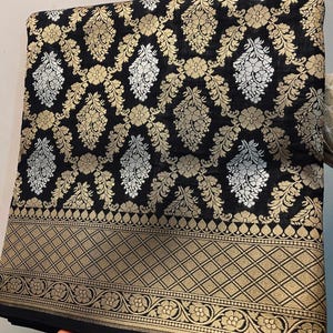 Puede incluir: Tela negra con un estampado floral dorado y plateado. La tela tiene un borde dorado con un diseño geométrico. La tela está doblada y el tema general es textil.