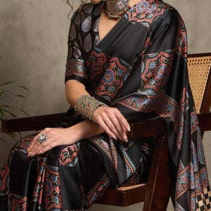 May include: A black sari with a detailed, colorful pattern featuring red, blue, and gold accents. The sari is draped over a person seated on a wooden chair. The sleeves and border of the sari showcase intricate designs.