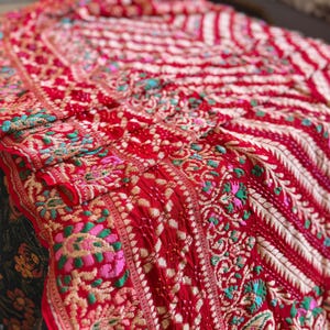 Puede incluir: Textil rojo vibrante con bordados intrincados. La tela presenta patrones florales y geométricos detallados en tonos rosa, verde y dorado. El diseño es rico y ornamentado, con una apariencia texturizada.