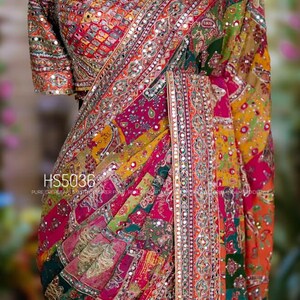 May include: A vibrant, multi-colored saree with intricate embroidery and mirror work. The design features a patchwork pattern in shades of pink, orange, green, and yellow. The text "HS5036" is visible on the fabric. A beaded necklace is also visible.